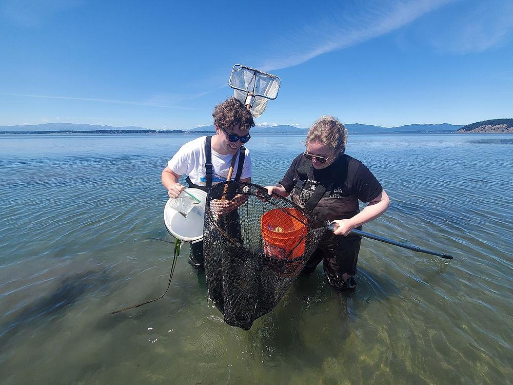 Two students stand in water holding net and research gear.